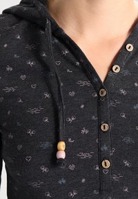 Mörkgrå huvtröja med små rosa och blå doodle-mönster, träknappar och dragsnören med trä- och rosa pärlor.