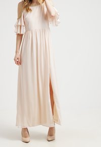 Vestido maxi de tela suave en color rosa claro, con diseño de hombros al aire, mangas con volantes y una abertura en el muslo. Detalles en acabado dorado.