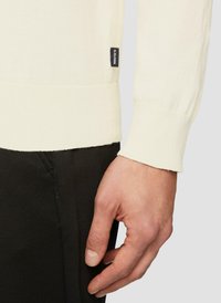 Licht crèmekleurig gebreid sweater met geribbelde manchetten en onderrand, met een zwart label aan de linkerkant. Draagt over donkere broek, met handdetail zichtbaar.