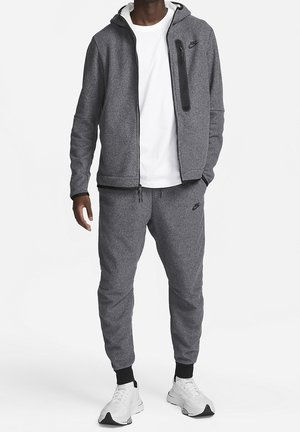 Man wearing gray Nike hoodie and matching joggers with white sneakers and white T-shirt, standing with one hand in pocket.