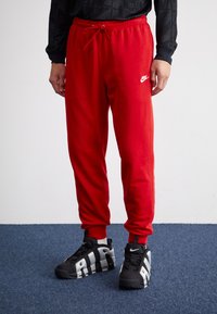Červené Nike tepláky s páskem na stažení v pase a bílým logem na levém stehně, doplněné černými sportovními botami s šedými akcenty.