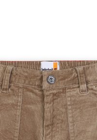 Brune corduroy bukser med en tekstureret overflade, en frontlukning med knap og et synligt Timberland-mærke til brandidentifikation.