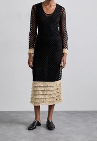 Donna che indossa un vestito lungo nero a maniche lunghe con motivo in rete, con dettagli in frange beige sui polsini e sull'orlo, abbinato a scarpe nere senza lacci.