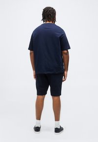 Άντρας με πλεγμένα μαλλιά φορά μπλε σκούρο φαρδιά μπλούζα και ταιριαστό σορτς, λευκές κάλτσες και μαύρα αθλητικά παπούτσια, γυρισμένος πλάτη σε απλό φόντο.