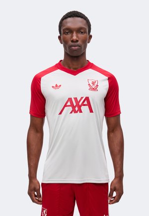 Homme portant un maillot blanc et rouge du Liverpool Football Club et un short rouge, debout face à l’avant avec une expression neutre.