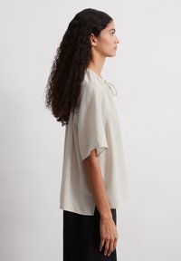 Beige Bluse mit kurzen Ärmeln und lässigem Schnitt, strukturiertem Stoff, runder Halslinie und Kordelzugdetail. Das Model trägt einen schwarzen Rock.