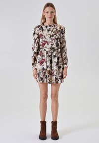 Bloemendruk jurk met lange mouwen, een ceintuur in de taille en een gelaagde rok. Kleuren zijn crème, bordeauxrood en donkergroen. Draagt met bruine enkellaarsjes.