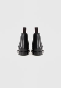 Par de botas de piel negra hasta el tobillo con tacones apilados y tiradores negros, presentando una marca en rojo sobre un fondo blanco.