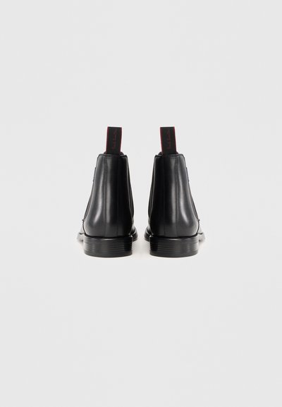 Par de botas de piel negra hasta el tobillo con tacones apilados y tiradores negros, presentando una marca en rojo sobre un fondo blanco.