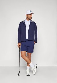 Man in marine blazer en shorts, witte polo, pet en sneakers, met een golfclub in zijn hand, staand met gekruiste benen tegen een plain achtergrond.