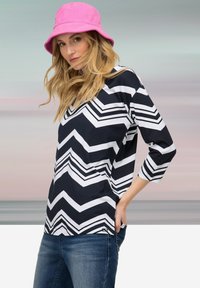 Top s námornicko modrým a bílým šikmým vzorem, se třičtvrtě rukávy, v kombinaci s modrými džínami a růžovým kordovým kloboukem.