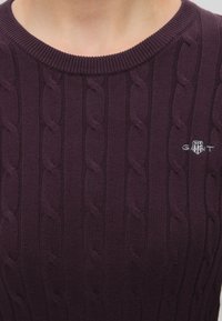 Gros plan d'une personne portant un pull en maille torsadée violet foncé avec un col rond et un petit logo brodé GANT sur la poitrine.