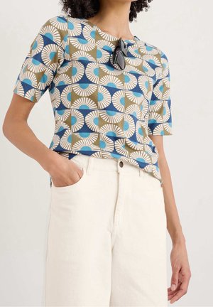 Žena v modrém, bílém a olivovém tričku s geometrickým vzorem a krátkým rukávem zastrčeném do vysokých bílých kalhot, s brýlemi zavěšenými na límci.