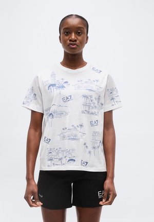 Femeie cu părul împletit, purtând un tricou alb cu imprimeu grafic albastru și pantaloni scurți negri, stând în fața unui fundal alb simplu.