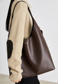 Brown leather tote bag with a spacious design. Features a smooth texture and a subtle gold logo. Paired with a cream knit sweater.