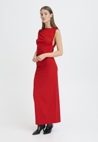 Mujer con vestido maxi rojo sin mangas con aberturas laterales y botines negros, de pie contra un fondo blanco liso.