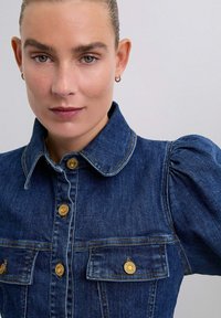 Mørk denimjakke med krave, guldknapper og puffede ærmer. Har to brystlommer og syningsdetaljer.