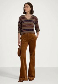 Γυναίκα ντυμένη με ένα ριγέ πουλόβερ V-neck, καφέ φλαθαρισμένα παντελόνια από κόκκινο ύφασμα, κρατώντας μια μικρή καφέ τσάντα στον παράλληλο φόντο.