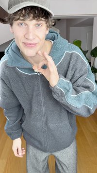 Graue Fleecejacke mit blauen Akzenten und weißer Einfassung. Das Modell trägt graue gestreifte Hosen. Kurze lockige Haare und eine Mütze, macht die Geste "OK".