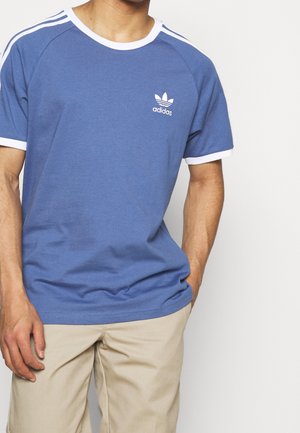 Mann trägt ein blaues Adidas-T-Shirt mit weißem Besatz und Logo, kombiniert mit beigen Hosen, steht vor einem einfarbigen hellen Hintergrund.