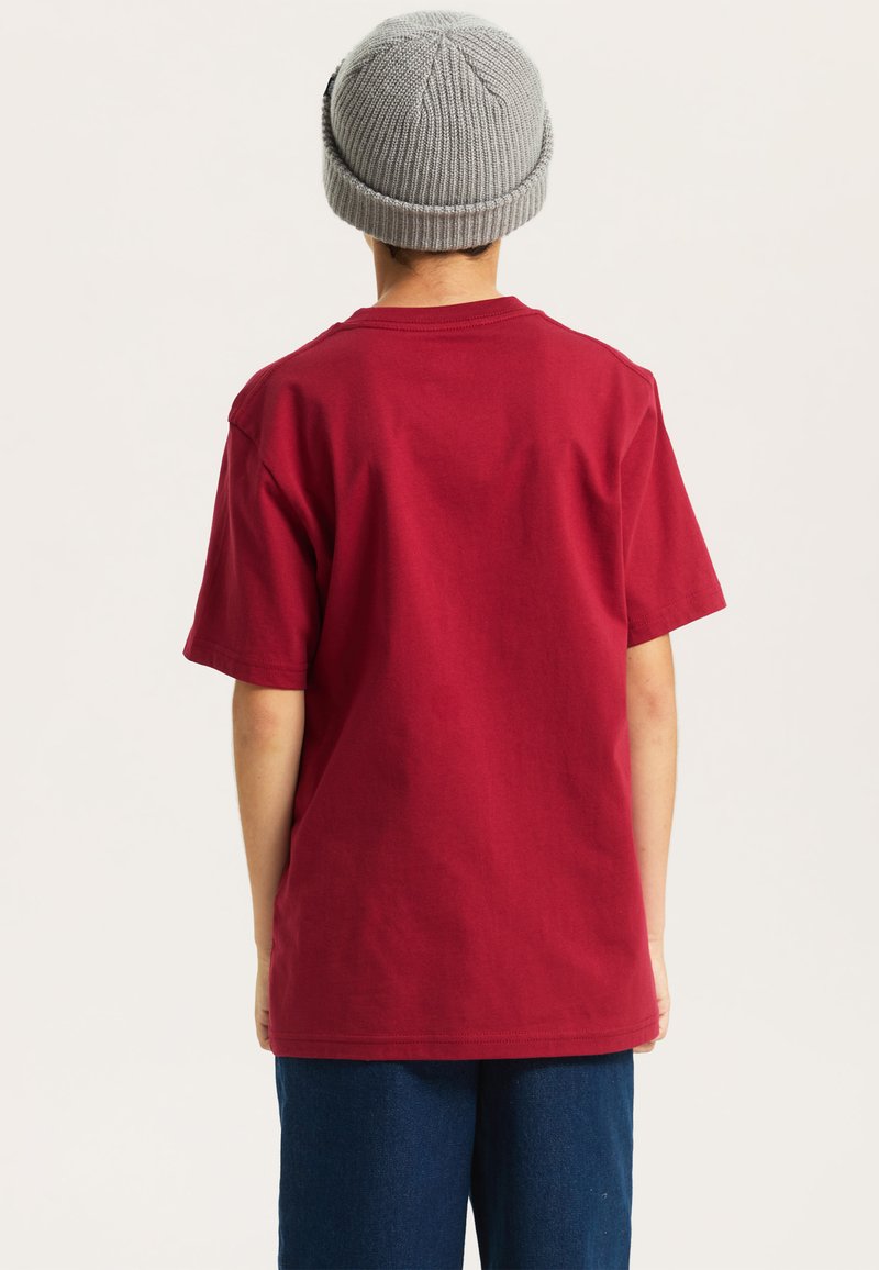 Czerwona bawełniana koszulka z okrągłym dekoltem i krótkimi rękawami, w połączeniu z niebieskimi dżinsami. Na głowie znajduje się szara dzianinowa czapka.