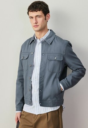 Young man with curly dark hair wearing a blue buttoned jacket over a striped white shirt and brown pants against plain background.