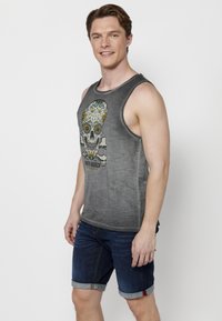 Szürke ujjatlan felső koponya grafikával, színes kiegészítőkkel, sötét farmer rövidnadrággal párosítva, amelynek az alja fel van hajtva. Sima textúra, laza szabás.