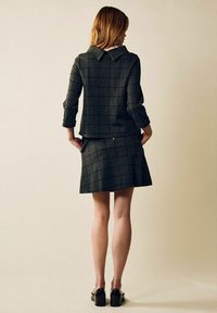 Kvinna som står med händerna i fickorna, klädd i en mörkgrå rutig kavaj och kjol, svarta skor med blockklack, vänd bort från kameran.