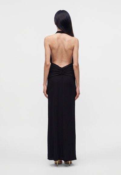 Woman with long dark hair wearing a floor-length black halter dress with an open back and gold high-heeled shoes, standing facing away.