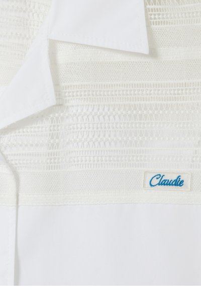 Blusa bianca con bordi in pizzo testurizzato, colletto classico e etichetta "Claudie" ricamata in blu nell'angolo in basso a destra.