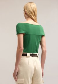 Zielony dopasowany top z krótkimi, nieco opadającymi rękawami; w połączeniu z beżowymi spodniami z wysokim stanem, zapinanymi na brązowy pasek.