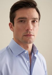 Camicia azzurra chiara con colletto strutturato, apertura frontale con bottoni e dettagli a fantasia all'interno del colletto. Il tessuto appare liscio e finemente tessuto.