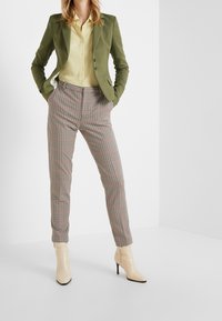 Blazer verde oliva sobre una camisa amarilla abotonada, combinada con pantalones de cuadros en rojo y beige, complementados con botines de tacón beige.
