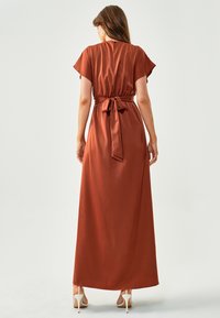 Dolga rjavkasta obleka iz gladkega materiala. Ima kratke, lepljive rokave in pas, ki ustvarja tekočo silhueto.