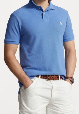 Man met een blauw poloshirt met korte mouwen, witte broek, een bruine gevlochten riem en een bruine leren horloge, met zijn handen in zijn zakken.