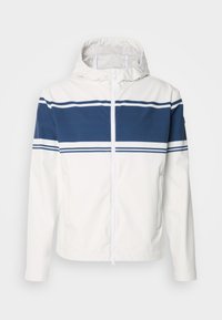 Hvid hættejakke med lynlås foran, der har en blå vandret stripe og tynde hvide kanter over brystet. Glat tekstur.