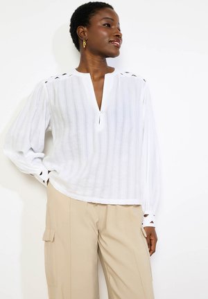 Žena v bielej blúzke s dlhými rukávmi s výrezmi a béžových cargo nohaviciach, stojaca s jednou rukou v vrecku, pozerá sa bokom a usmieva sa.