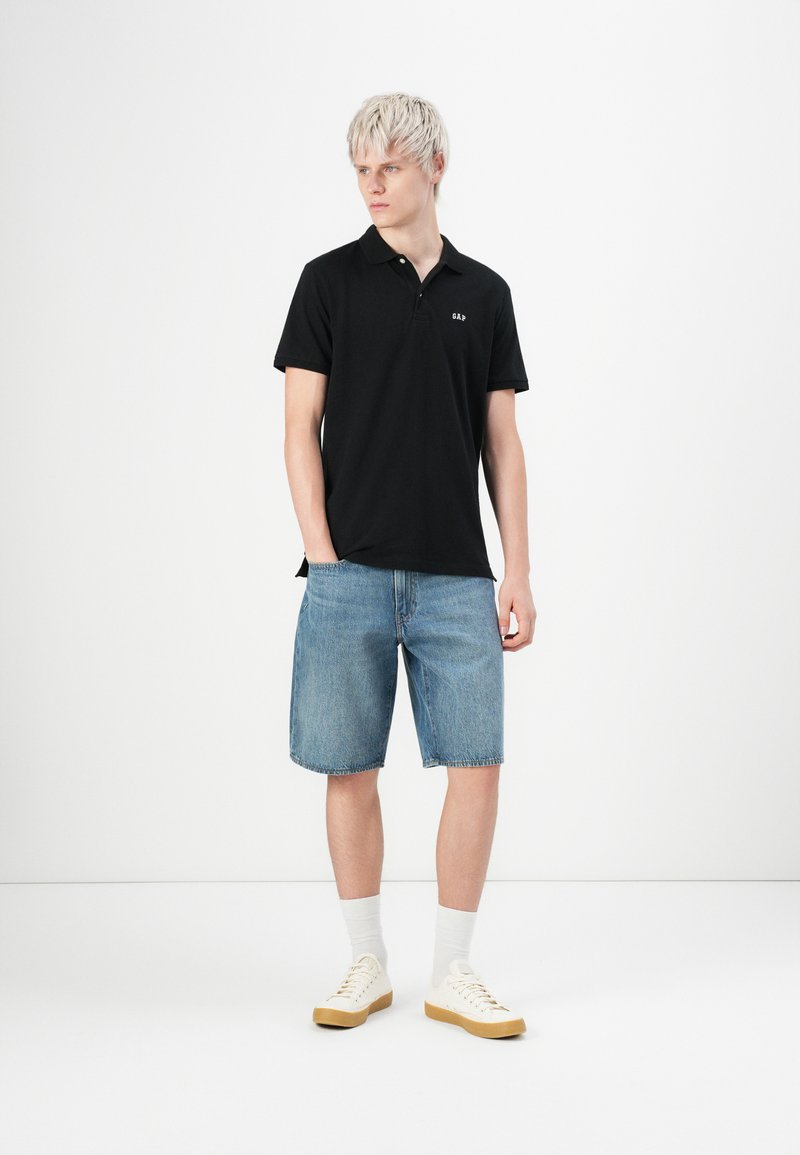 Polo noir avec col et logo sur le devant, associé à un short en denim bleu clair. Le modèle porte des chaussettes blanches et des baskets beiges.