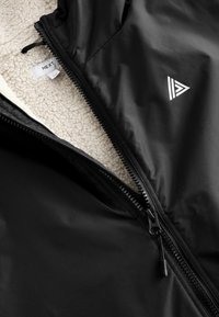 Zwart jack met een gladde buitenlaag en een crème, pluizige binnenvoering. Het heeft een ritssluiting en een driehoekig logo op de borst.