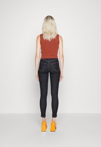 Jeans skinny de denim escuro com uma cintura alta, apresentando bolsos traseiros e costura em contraste, combinados com um top sem mangas de cor rust e sapatos de salto laranja.