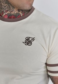 Crèmekleurig T-shirt met een ronde bruine halslijn en strepen op de mouwen. Kenmerkt zich door een donkerbruine geborduurde logo op de borst.