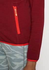 Manche de veste texturée bordeaux avec poche zippée rouge vif et bordure, associée à des leggings gris et blancs à motif abstrait.