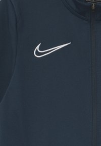 Chaqueta deportiva de color azul marino hecha de tela texturizada, que presenta un logotipo de Nike bordado en blanco en la parte superior izquierda y un cierre con cremallera.