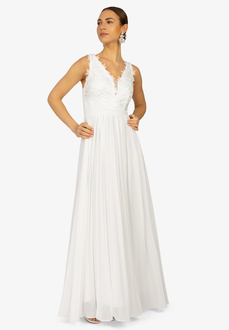 Robe blanche avec un décolleté en V, des détails en dentelle sur le corsage et une jupe plissée. Conception sans manches ; longueur au sol, texture de tissu douce.