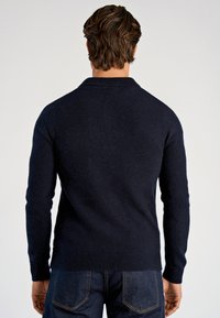 Granatowy, dopasowany sweter z dzianiny o fakturowanej powierzchni. Posiada prążkowany kołnierz i mankiety, zestawiony z ciemnymi jeansami.