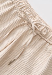 Gros plan sur un pantalon en tissu rayé beige avec une ceinture élastique et un cordon de serrage noué avec des embouts métalliques.