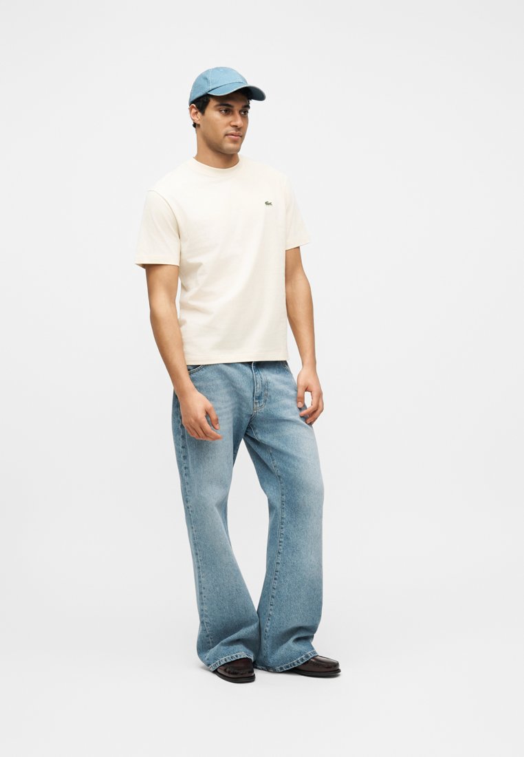 Homme debout portant une chemise à manches courtes beige clair, un jean bleu clair ample, des chaussures foncées et une casquette de baseball bleu clair, sur un fond uni.