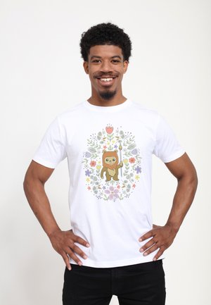 Smilende mand med kort krøllet hår iført en hvid T-shirt med en tegneserie bjørn, der holder en spyd omgivet af farverige blomster.