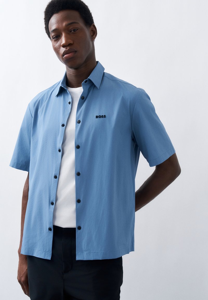 Νεαρός άνδρας που φοράει γαλάζιο πουκάμισο με κοντά μανίκια και κουμπιά πάνω από ένα λευκό μπλουζάκι, στέκεται μπροστά σε ένα απλό ανοιχτό φόντο.