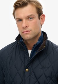 Giacca imbottita blu navy con colletto alto, accessori in oro e dettagli in corduroy a contrasto. Sotto, una giacca blu con zip.